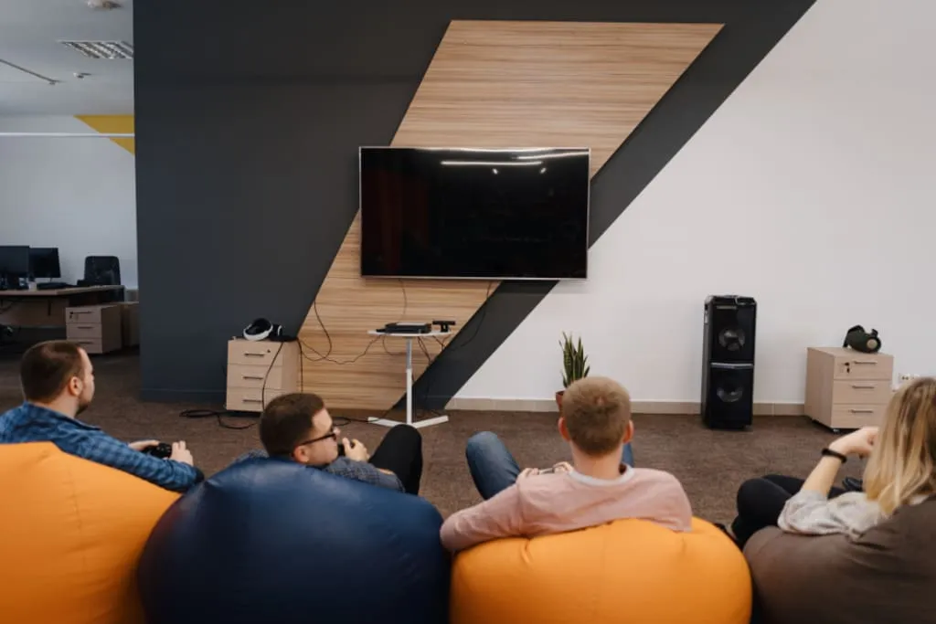 Group of coworkers relaxing on colorful bean bag chairs, playing video games together in front of a wall-mounted TV in a modern office lounge