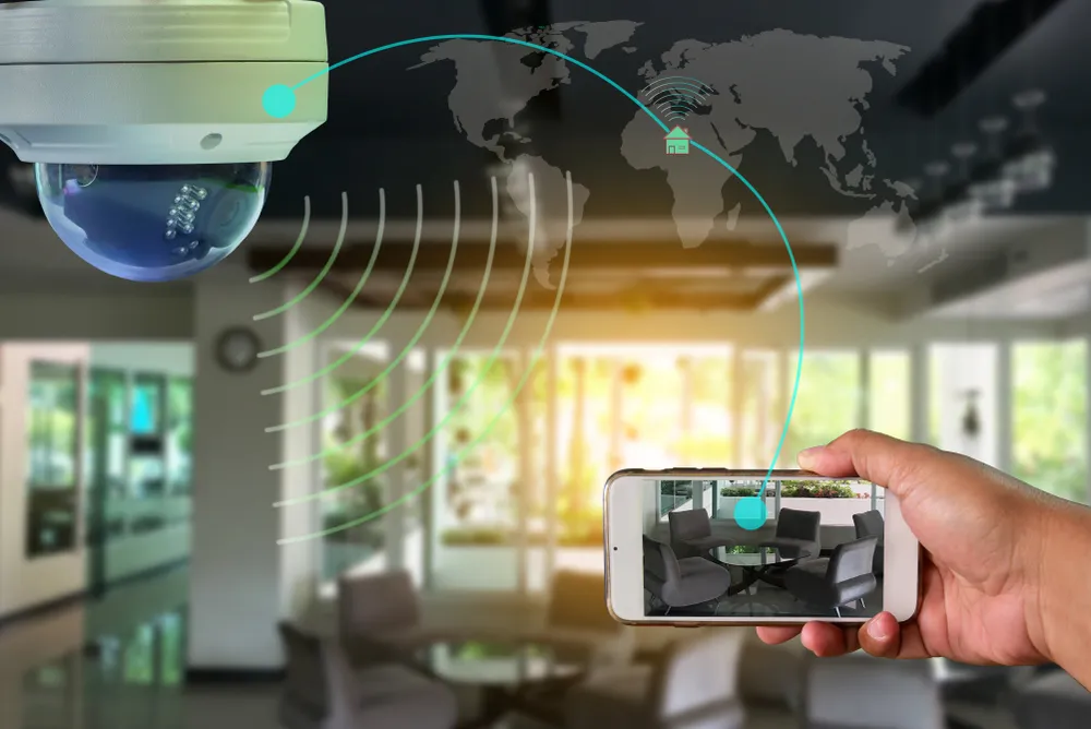 Ceiling-mounted security camera connected via a global signal graphic to a smartphone showing live remote room monitoring footage
